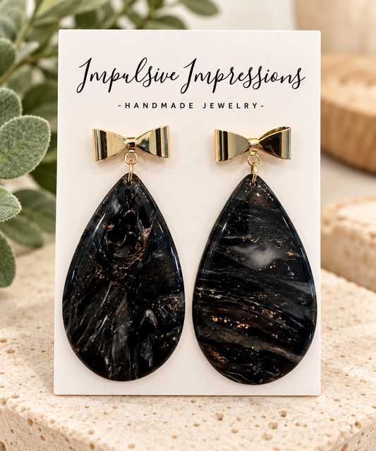 Black marbled teardrop earrings with gold bow studs on warm neutral boutique background
